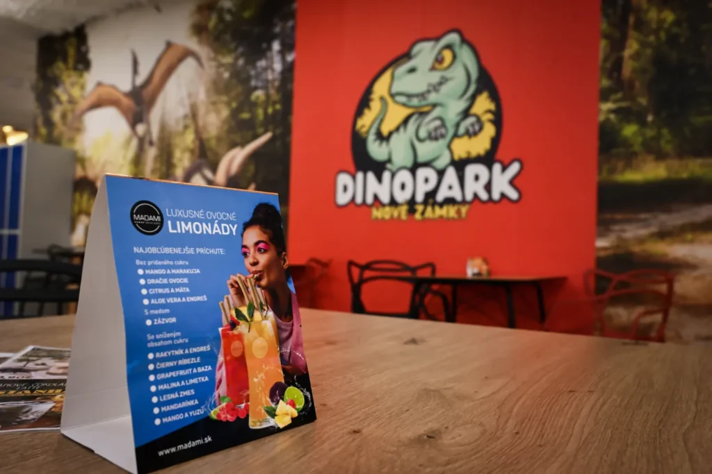 Informačný stojan pri recepcii s logom Dinopark Nové Zámky a tematickou výzdobou v pozadí.